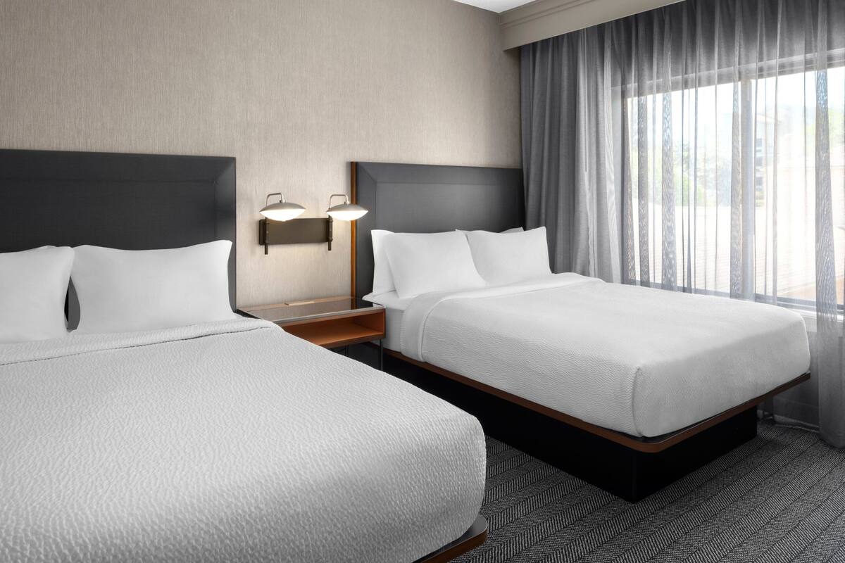 Een hotelkamer met twee bedden, een nachtkastje met een lamp en een groot raam met gordijnen.