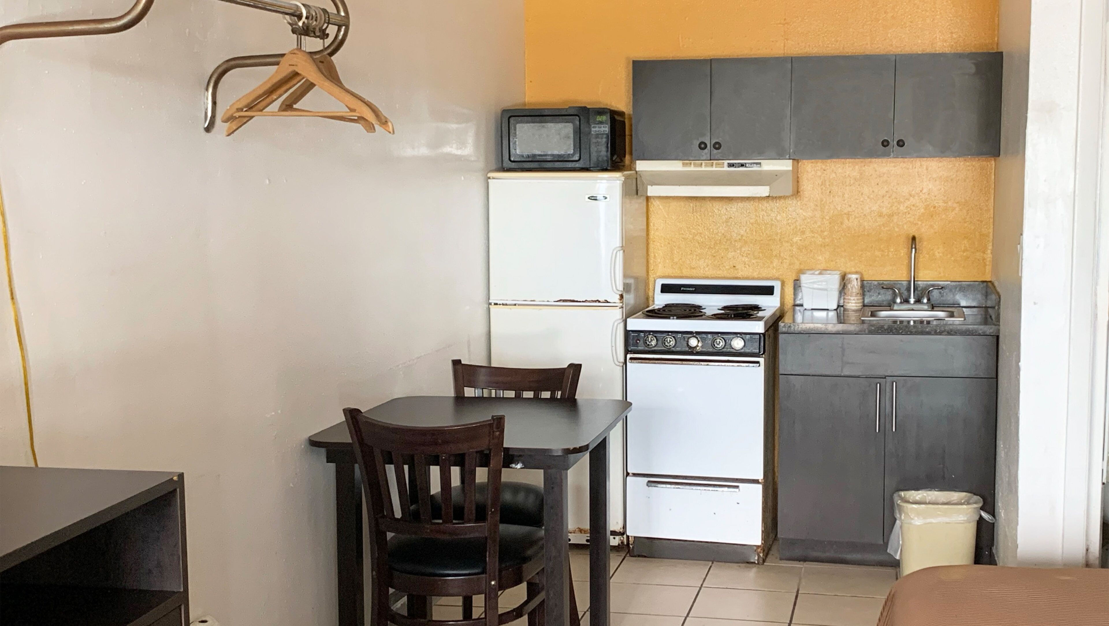 A compact kitchenette with a white refrigerator, a stove, and a microwave. There is a wooden chair and a small table.