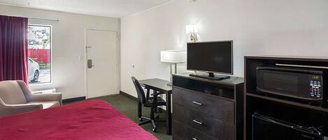 A hotel room with a bed, a desk with a computer, a chair, a microwave, and a window with curtains.