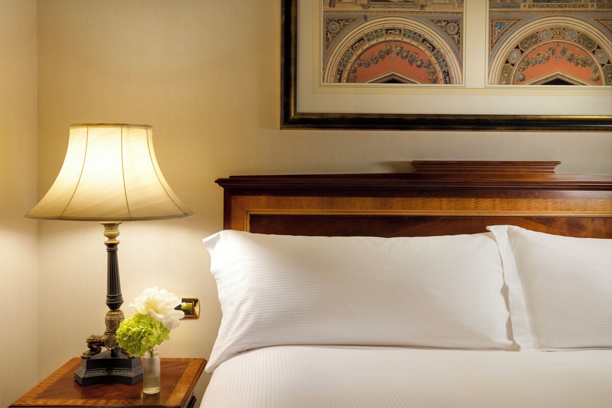 Una cama bien hecha con sábanas blancas, un cabecero de madera y una mesita de noche con una lámpara y un florero.