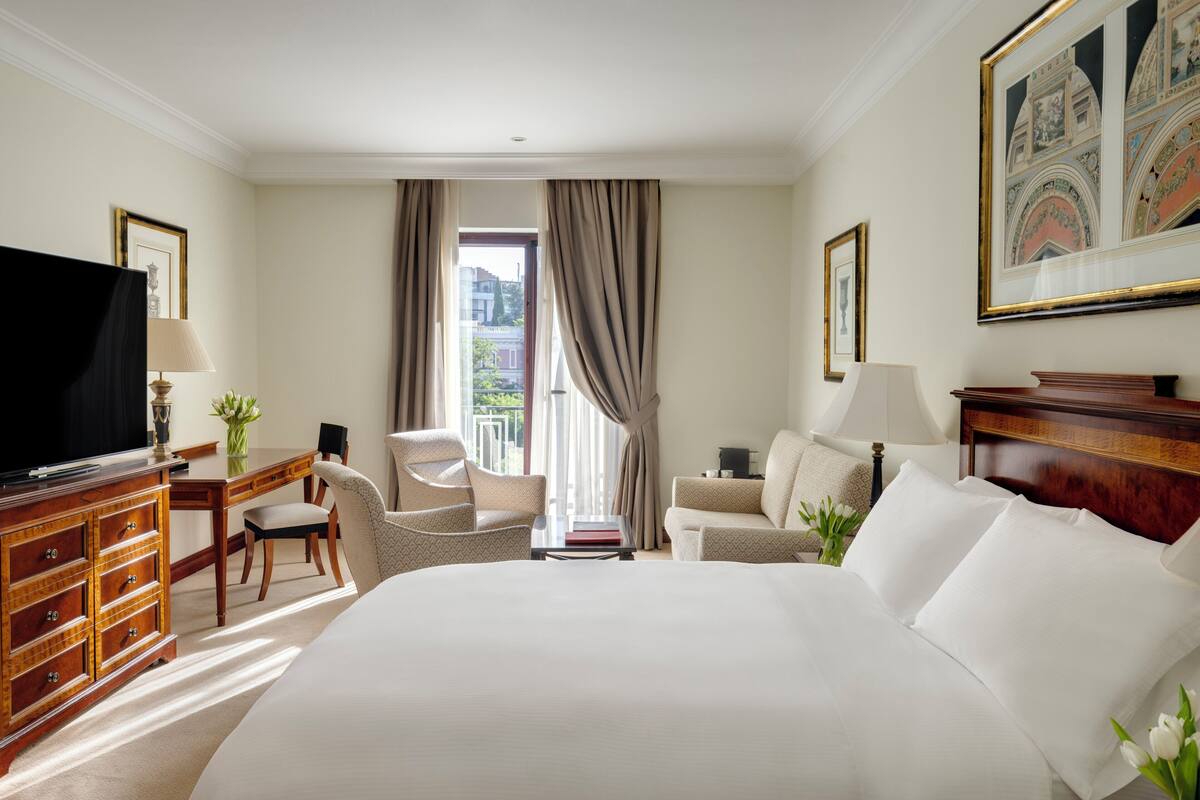 Una habitación de hotel con una cama grande, un escritorio con lámpara, un televisor y una ventana con cortinas.