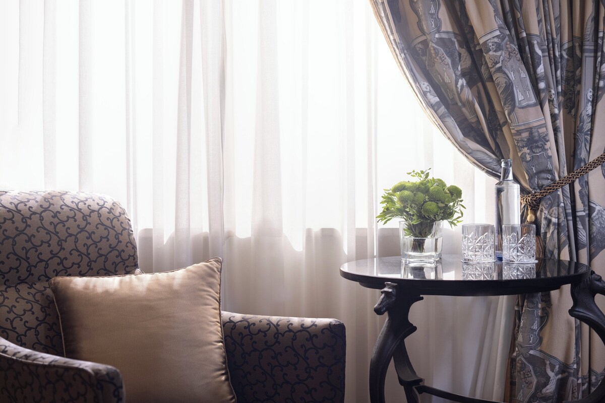 Una habitación con un sillón estampado, una mesita con un florero y vasos, y cortinas translúcidas.