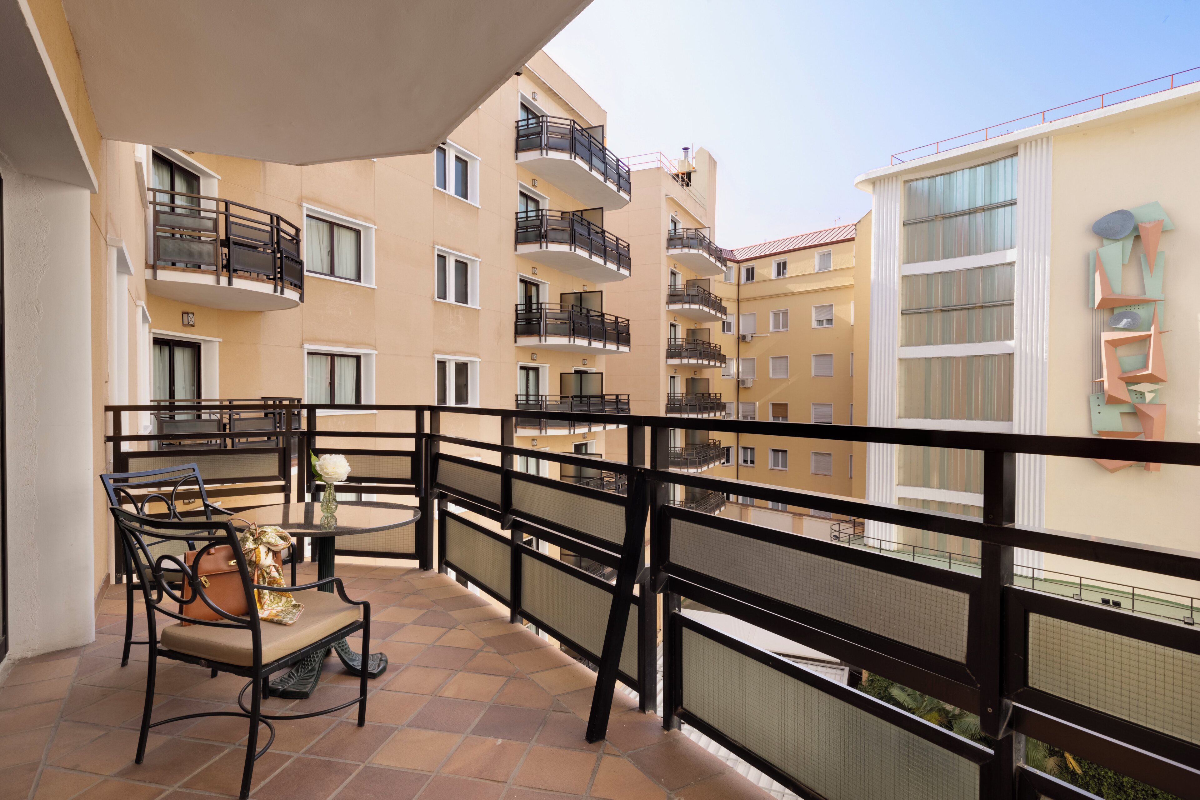 Un balcón con una silla y una mesita, con vistas a un edificio residencial con múltiples balcones y ventanas.