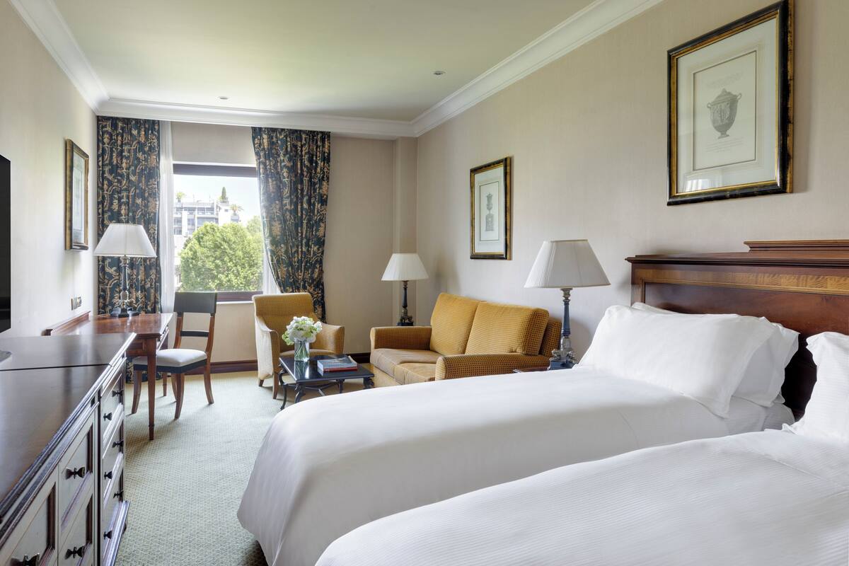 Habitación de hotel con dos camas, un escritorio, una silla, un sofá y una ventana con cortinas.