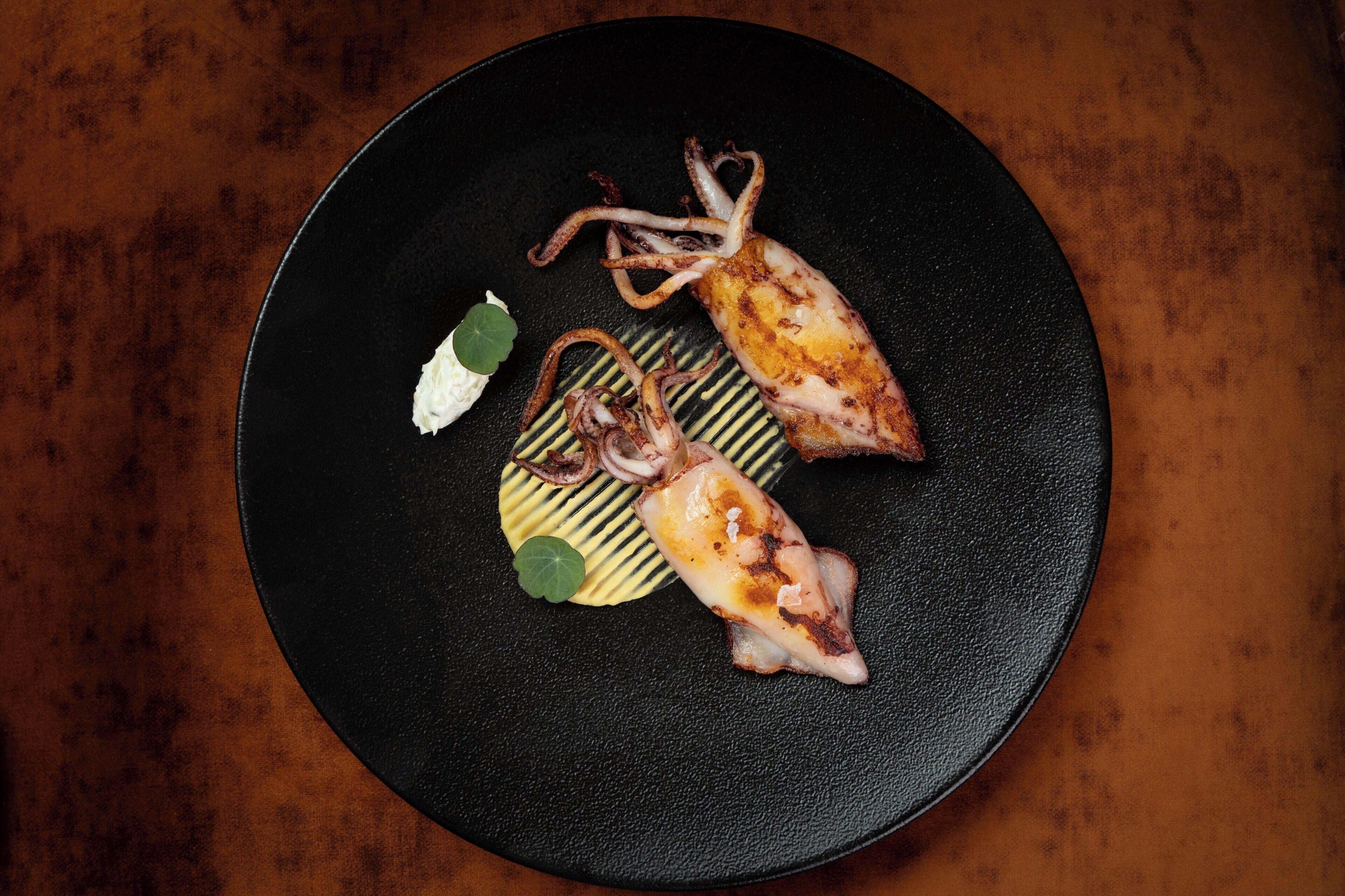 Une assiette noire avec des calmars grillés et un petit objet blanc sur une surface texturée.