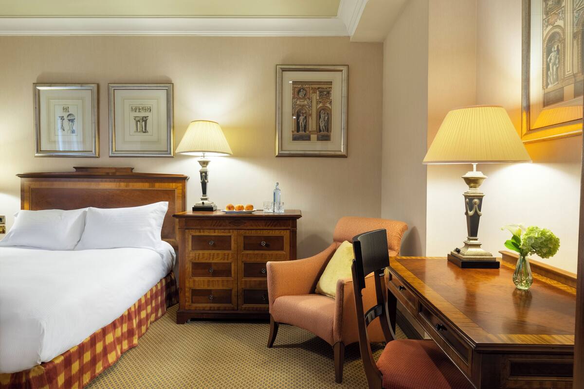 Habitación de hotel con cama, cómoda de madera, escritorio con lámpara y cuadros en la pared.