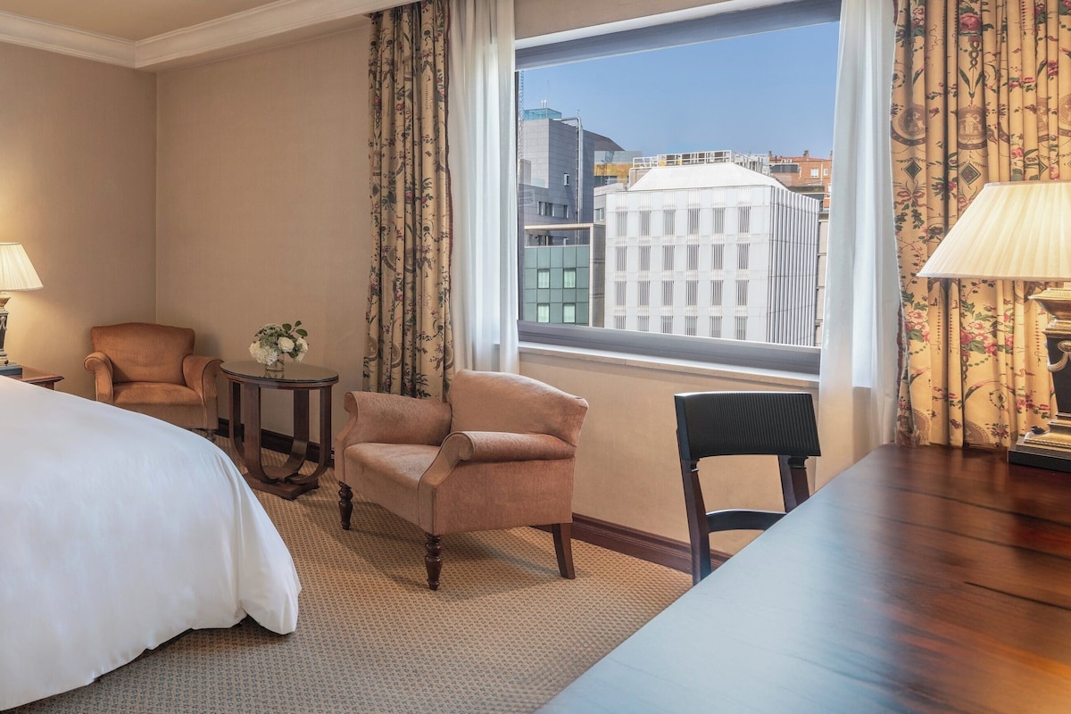 Habitació d'hotel amb un llit gran, dues làmpades de nit, una cadira, una taula petita amb un vas i vista a edificis per la finestra.
