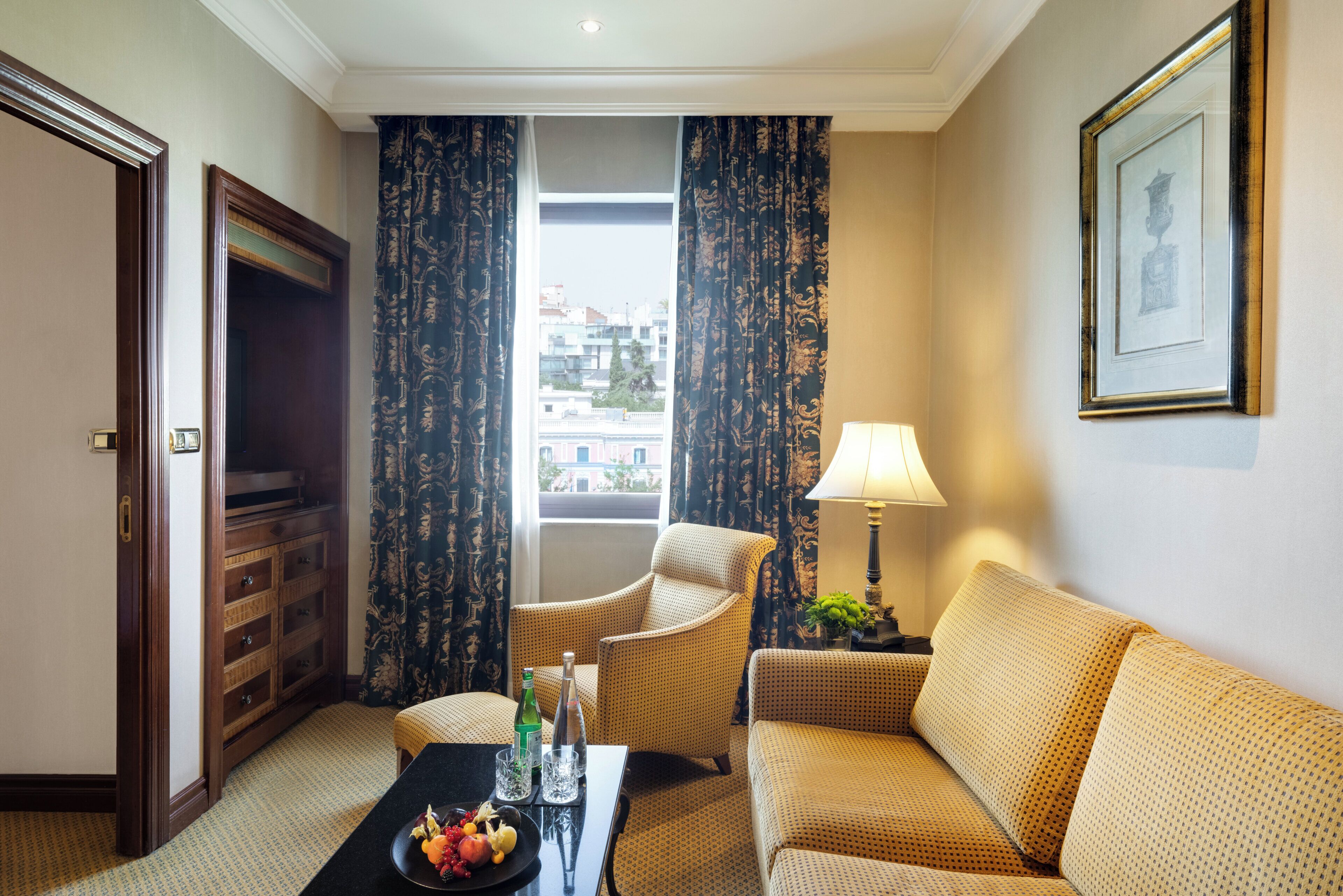 Habitación de hotel con sofá, sillón y una pequeña mesa con frutero y botellas.