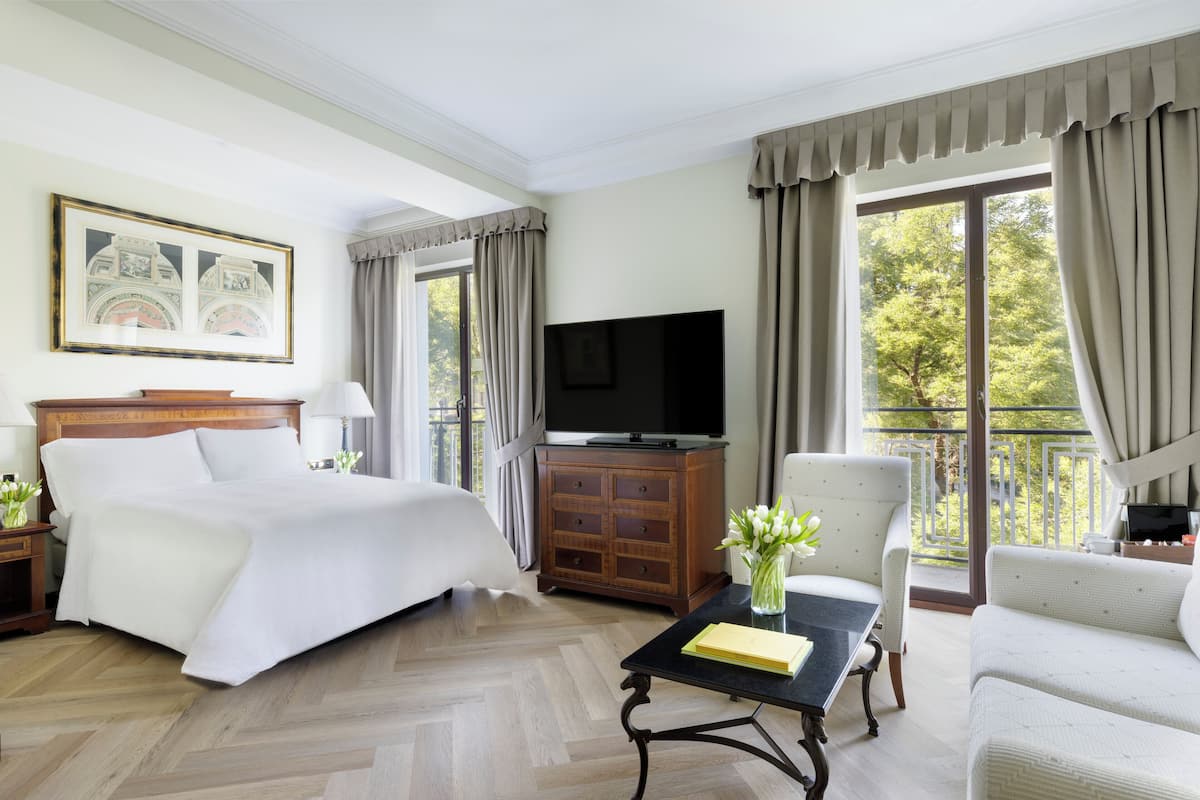 Habitación de hotel con una cama grande, un televisor de pantalla plana, un mueble de madera, una mesita con un jarrón de flores y un balcón con cortinas.