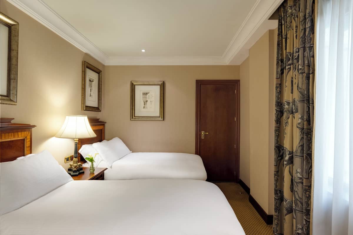 Habitación de hotel con dos camas, una mesita de noche con lámpara, cuadros en la pared y una puerta.