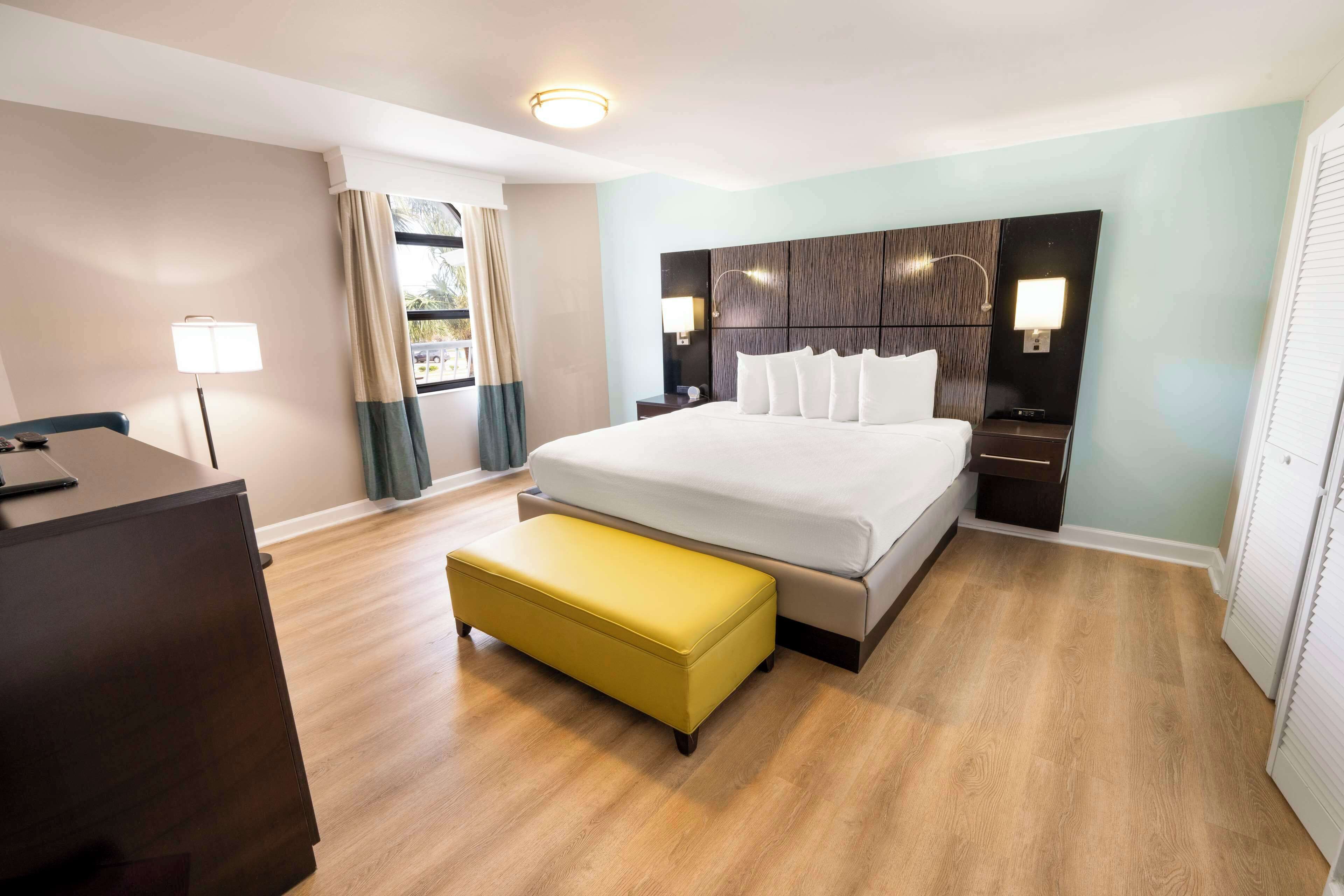 Habitación de hotel con cama, mesitas de noche, escritorio, banco y ventana con cortinas.