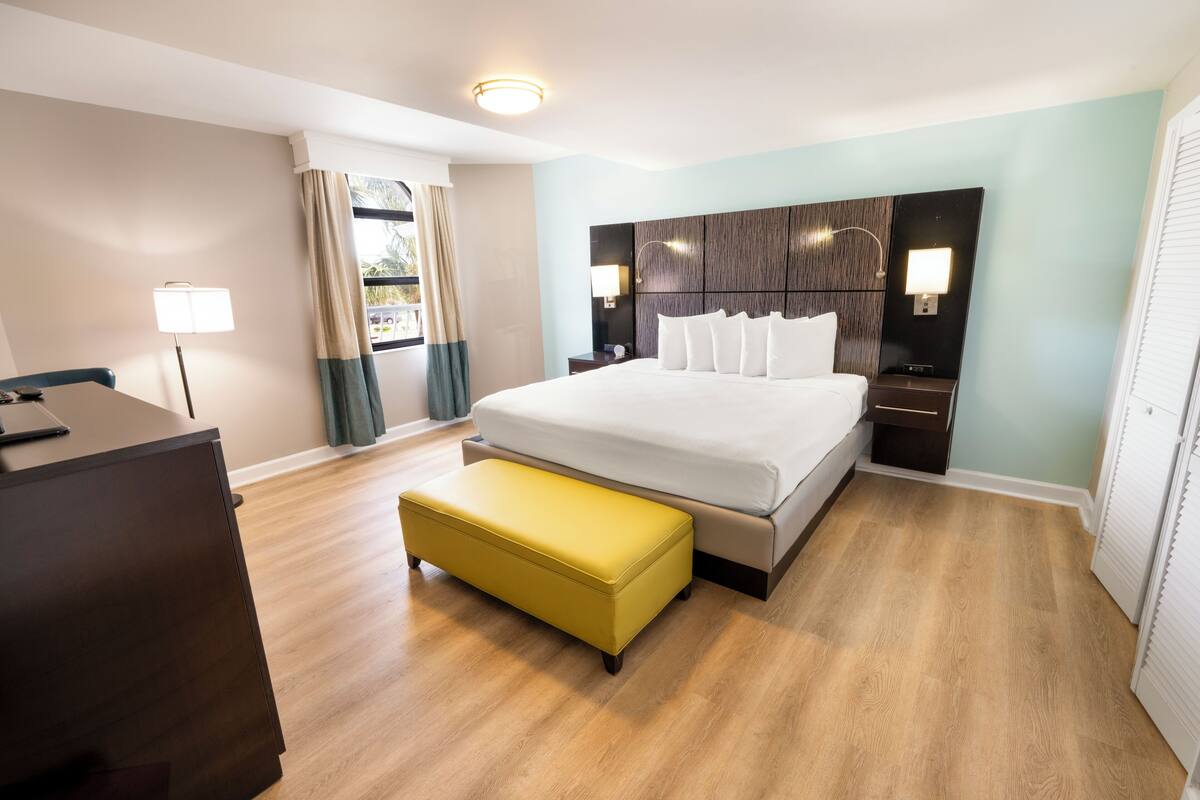 Quarto de hotel com cama, mesas de cabeceira, escrivaninha, cadeira e janela com cortinas.
