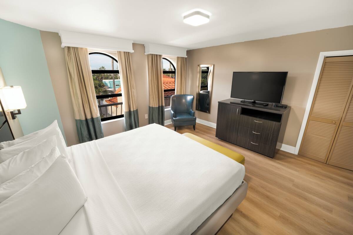 Quarto de hotel com cama, televisão, cadeira, escrivaninha e guarda-roupa.
