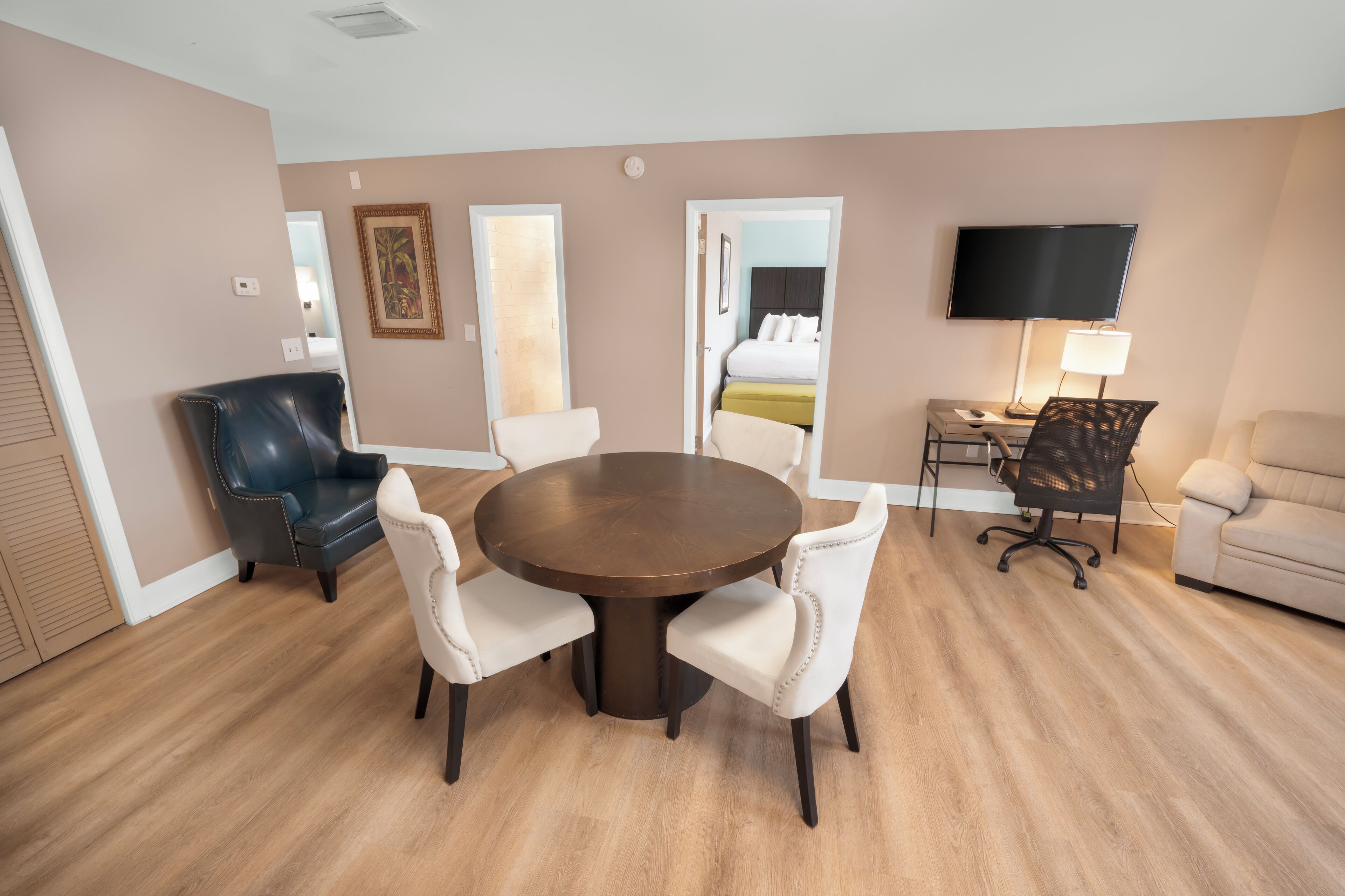 Habitación de hotel con una mesa de comedor redonda, dos sillas blancas, un sillón de cuero negro, un sofá, un televisor y una mesita de noche con lámpara.