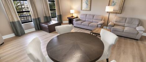 A living room with a round table, white chairs, a sofa, and a loveseat. There is a window with curtains, a lamp, and two framed pictures on the wall.