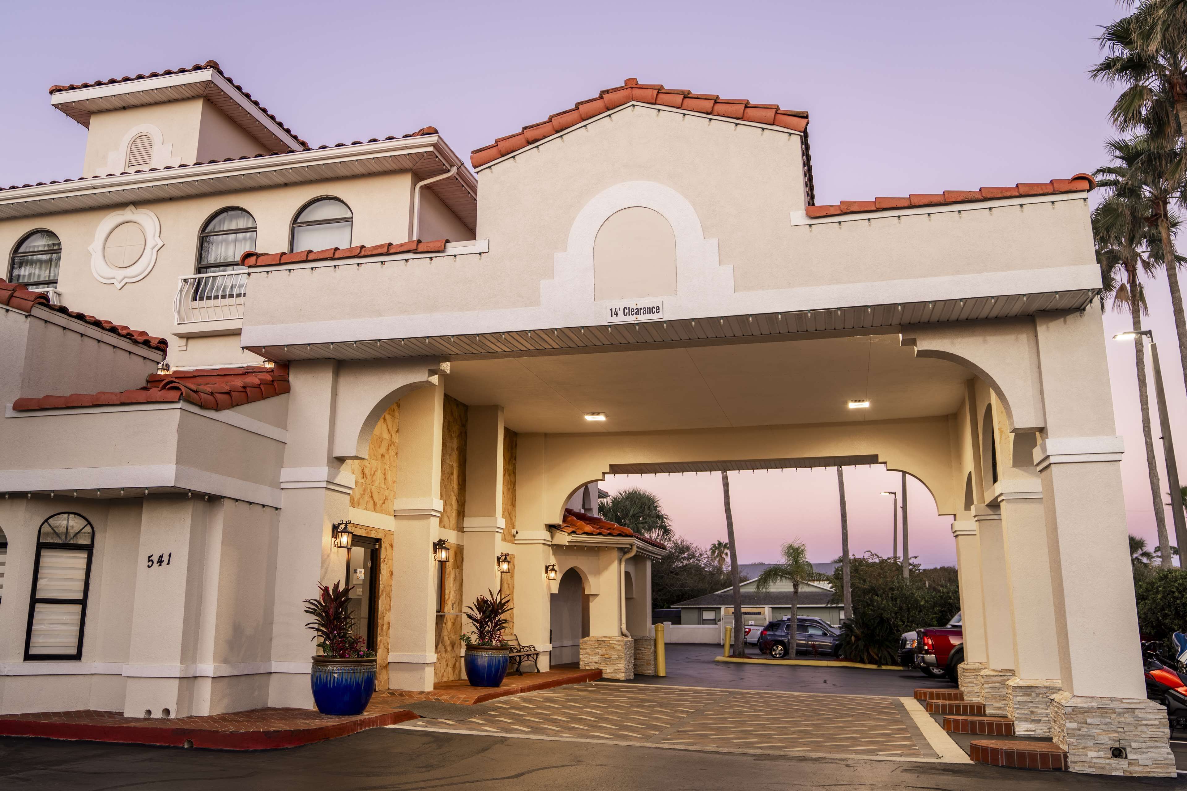 A hotel entrance with a covered driveway, a large potted plant, and a clear sky.
