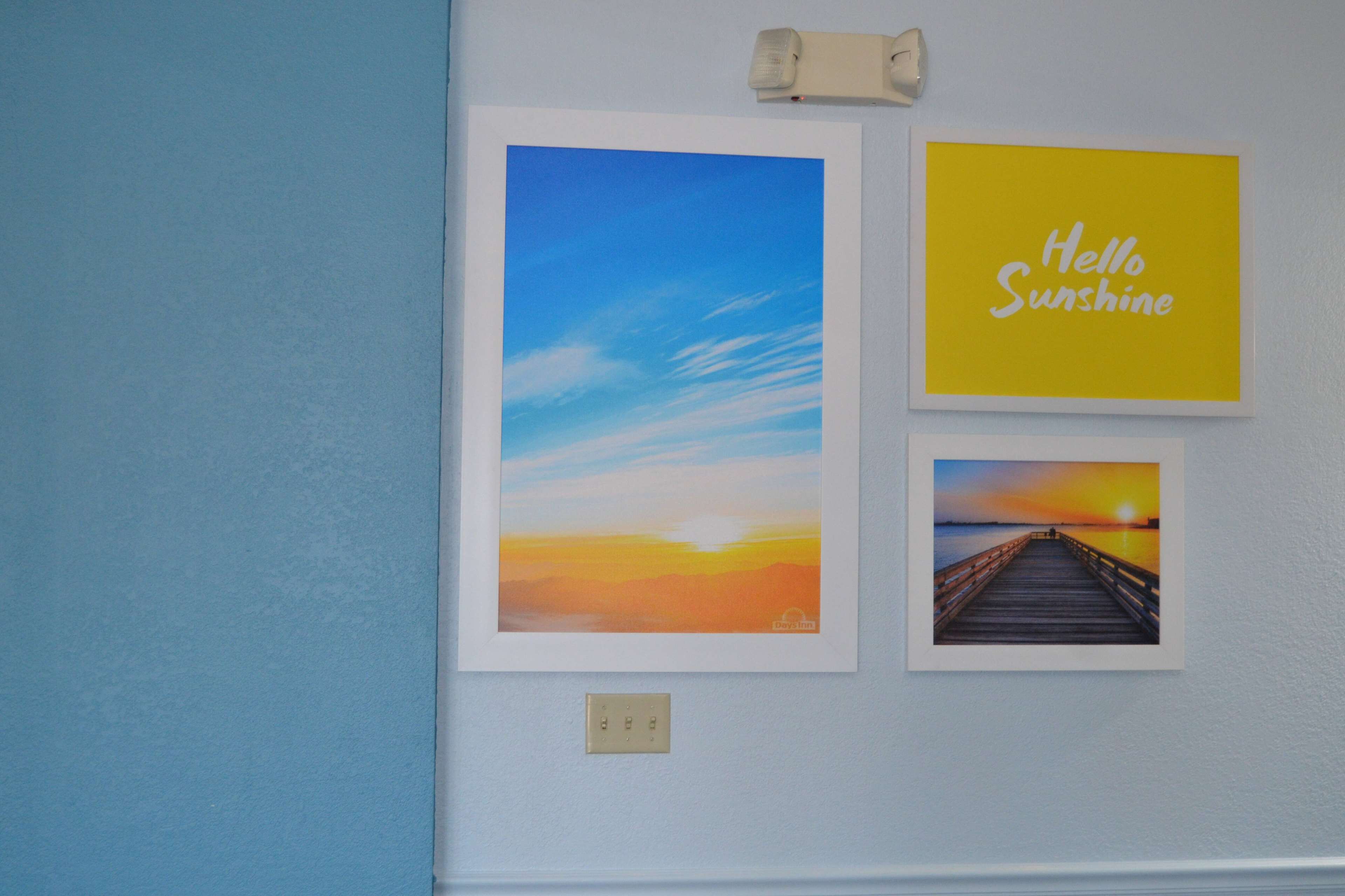 A wall with three framed pictures: a sunset, a 'Hello Sunshine' sign, and a pier.