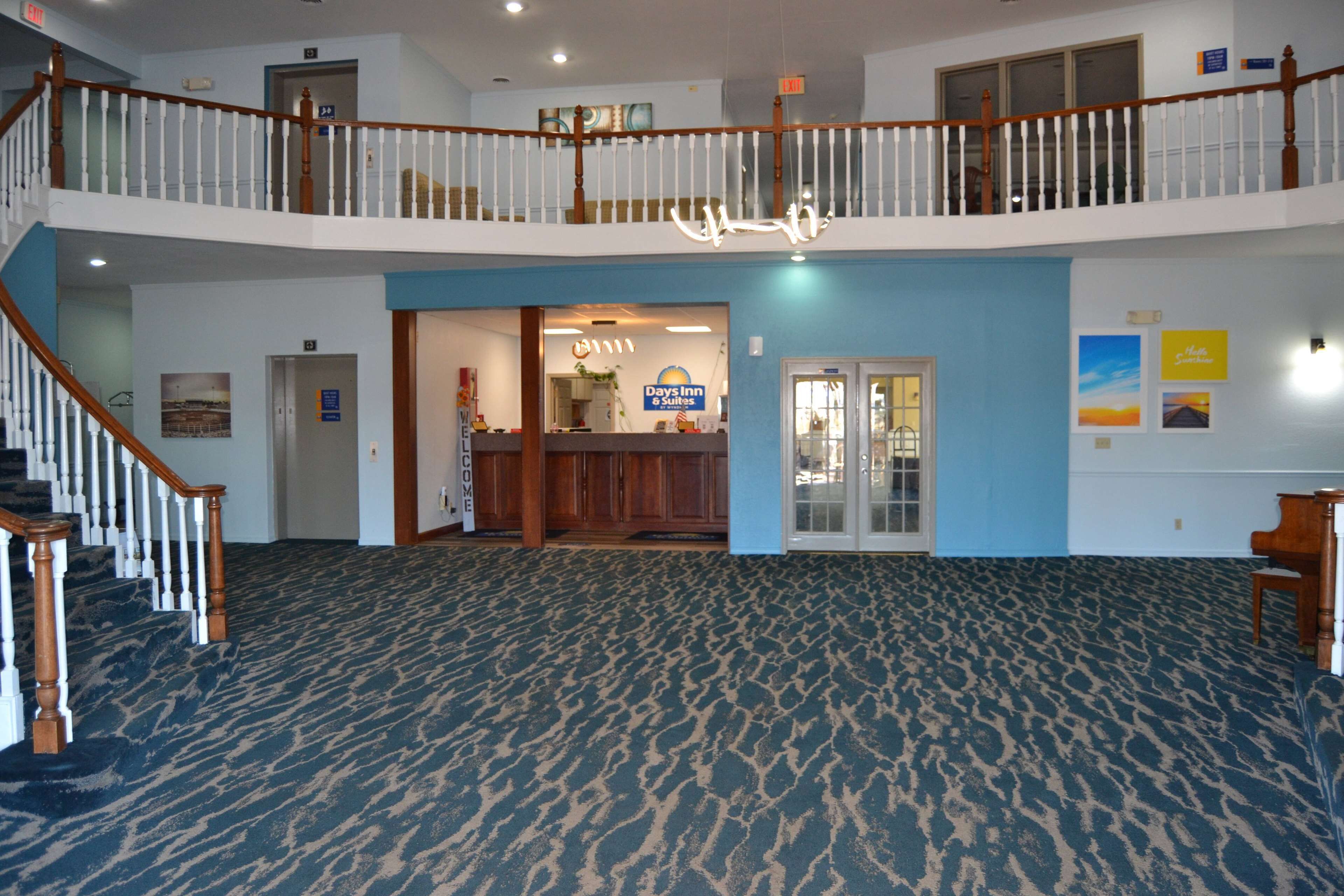 A hotel lobby with a blue carpet, wooden staircase, reception desk, and a balcony with a railing.