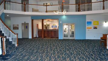 A hotel lobby with a blue carpet, wooden staircase, reception desk, and a balcony with a railing.