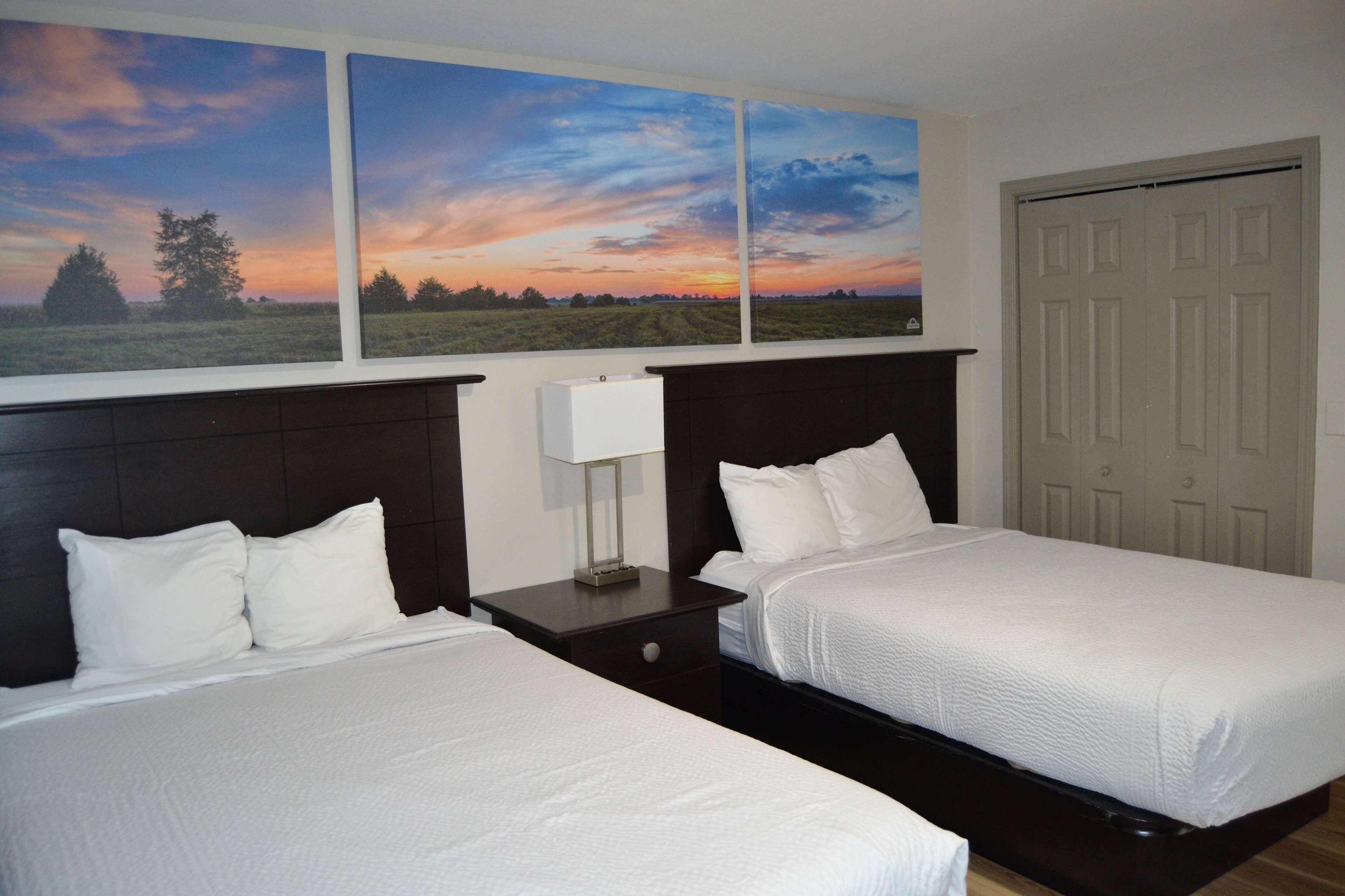 A hotel room with two beds, a nightstand with a lamp, and a large window with a sunset view.