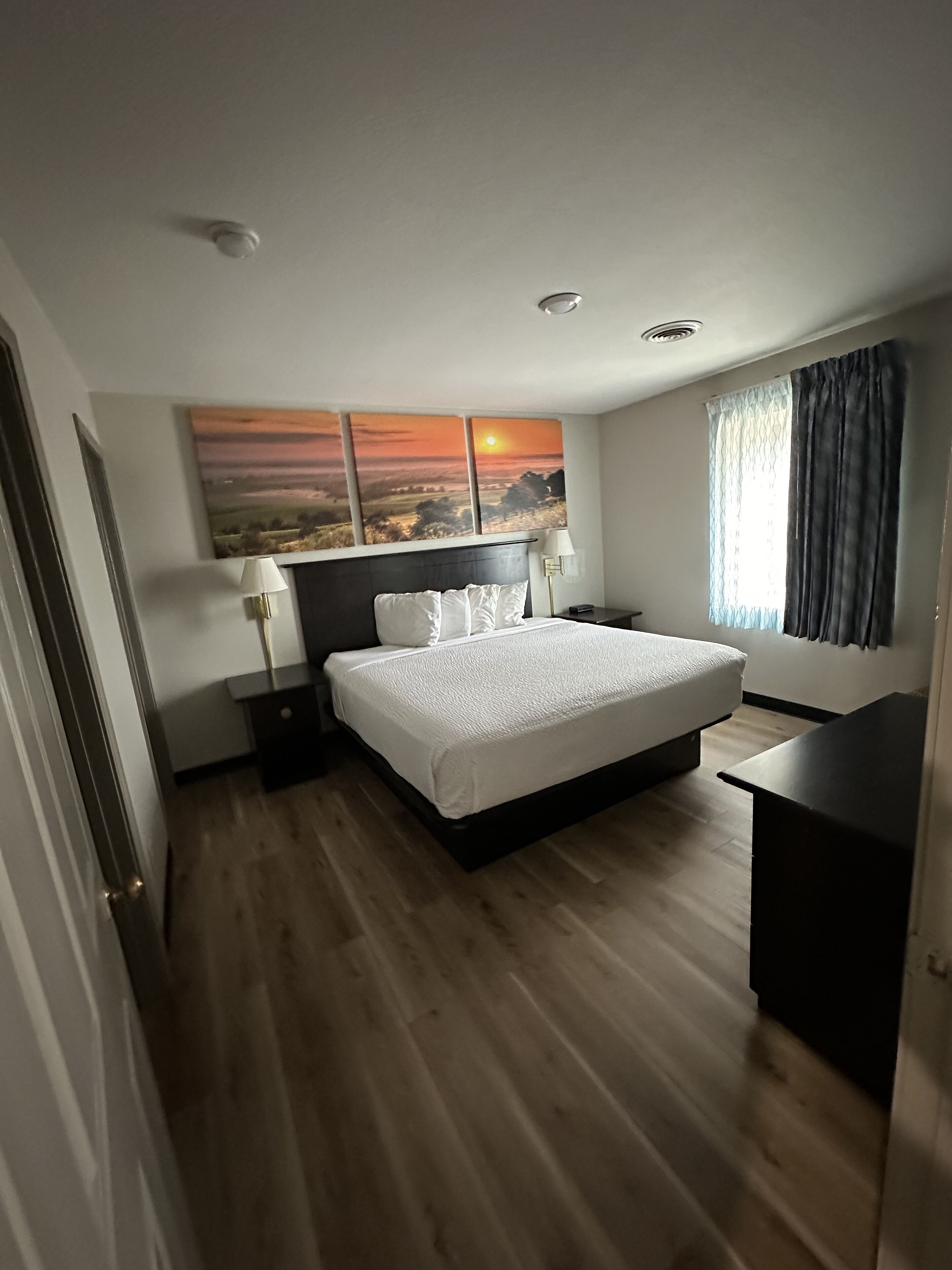 Ein Hotelzimmer mit Bett, Nachttischen, Schreibtisch, Lampe, einem Gemälde und einem Fenster mit Vorhängen.