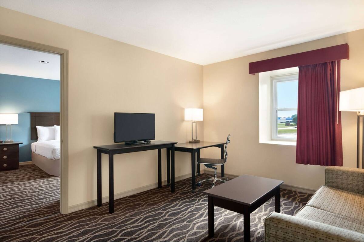 Habitación de hotel con cama, televisor sobre un escritorio, una silla, una mesita y una ventana con cortinas.