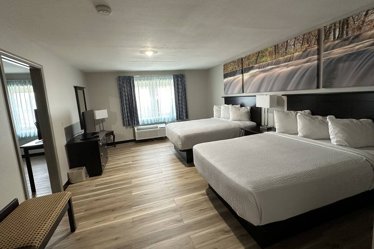 Habitación de hotel con dos camas, un escritorio, una silla, un televisor y un ventanal grande con cortinas.