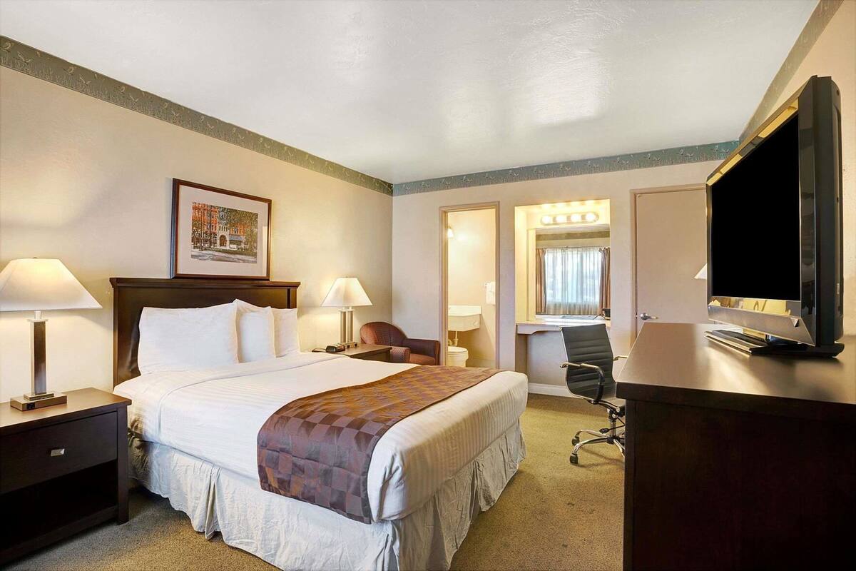Habitación de hotel con una cama, dos mesitas de noche con lámparas, un escritorio con una silla, un televisor y un espejo grande.