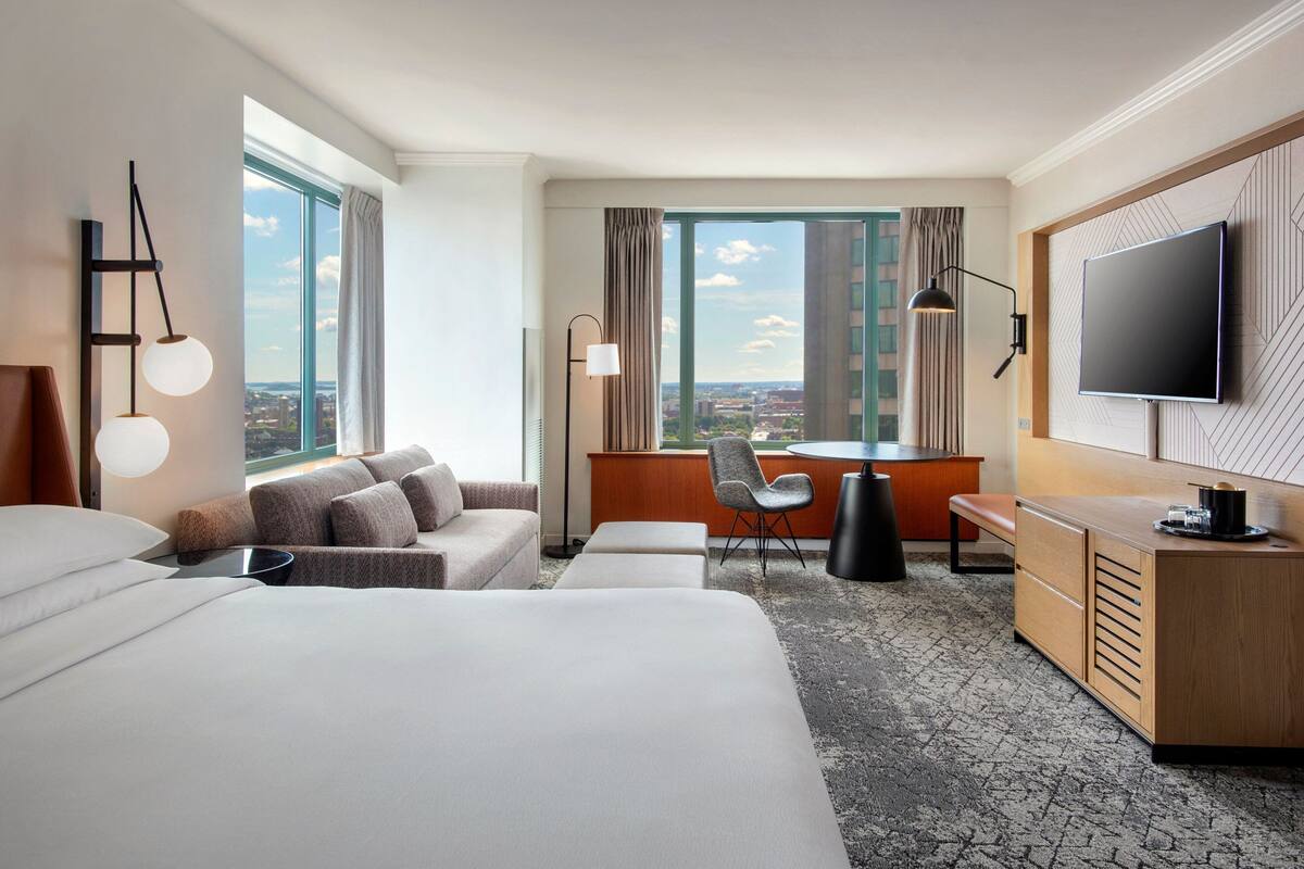 Camera d'albergo con un grande letto, una scrivania con una sedia, una TV e vista sulla città attraverso ampie finestre.