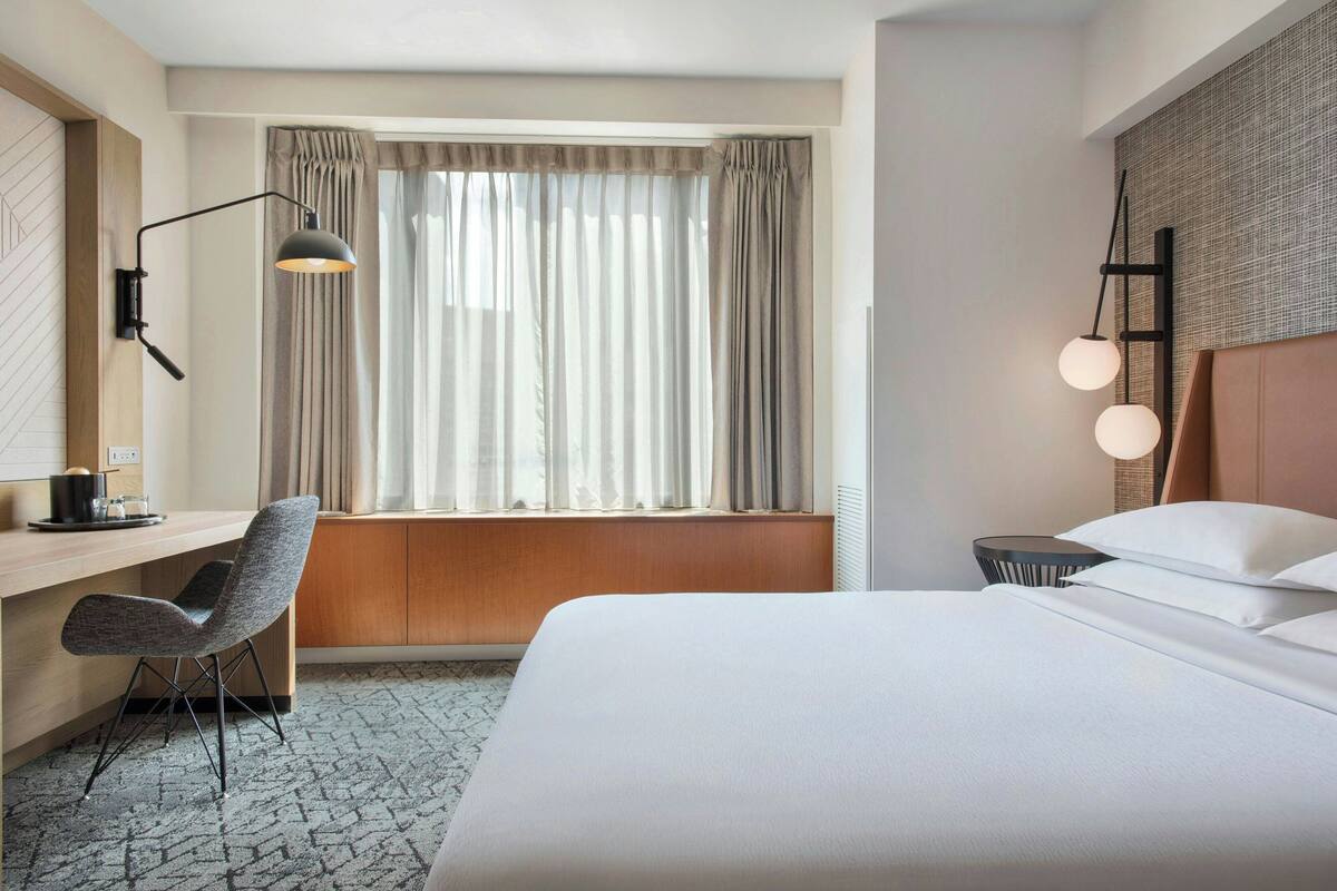 Una camera d'albergo con un letto, una scrivania, una sedia, una finestra con tende, una lampada a parete e un comodino con lampada.