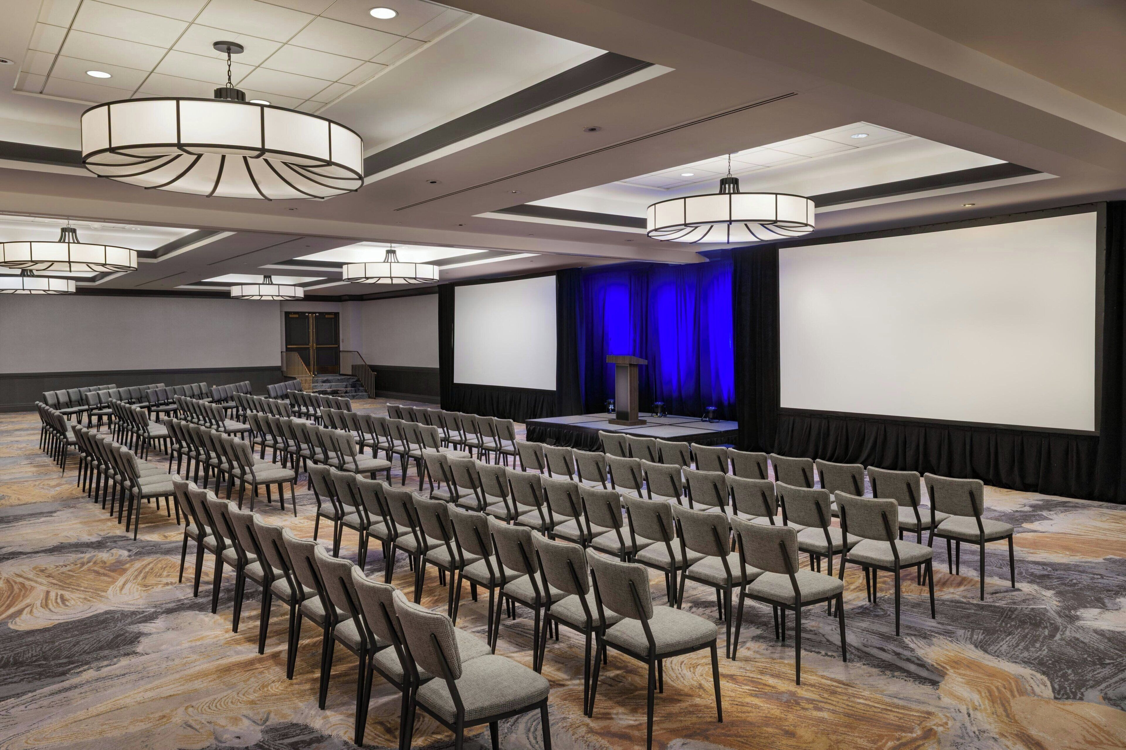 Un salón de conferencias con filas de sillas, un escenario con cortina azul y una pantalla grande.