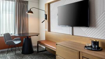 A modern hotel room with a flat-screen TV mounted on the wall, a wooden desk with a bench, a chair, and a small table with a tray.