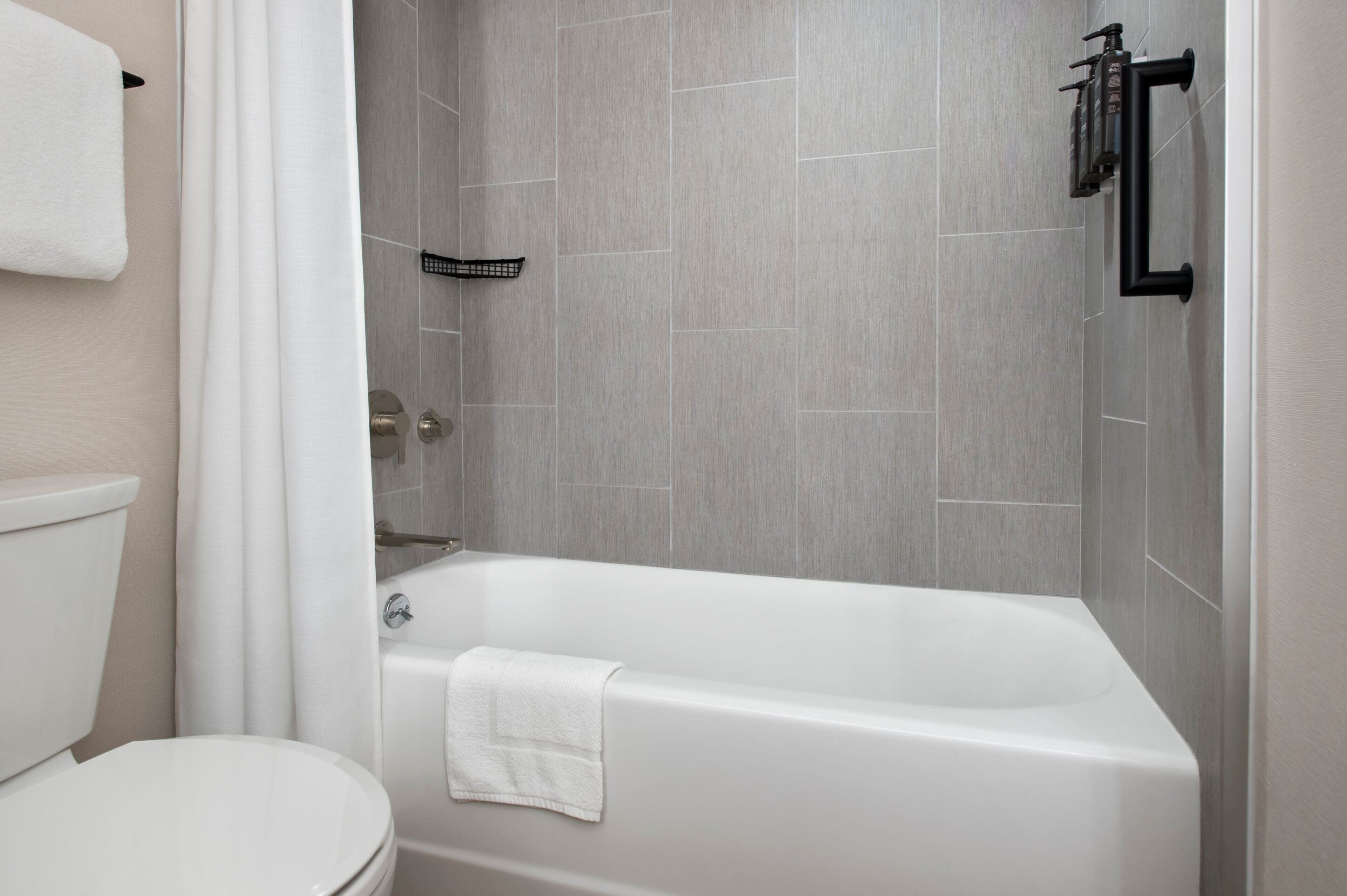 Un baño moderno con inodoro blanco, bañera con toalla blanca y paredes revestidas de baldosas grises.