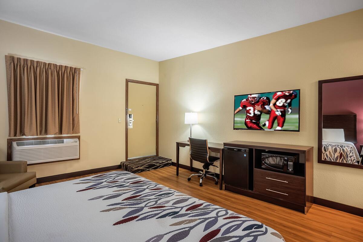 A hotel room with a bed, a desk, a chair, a TV, a microwave, and a mirror.