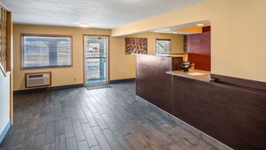 A hotel lobby with a reception desk, a large window with blinds, a door leading to a hallway, and a wall-mounted air conditioning unit. - Red Roof Inn Des Moines (Des Moines)