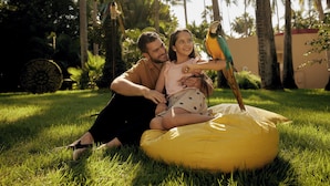 Un hombre y un niño sentados en un sillón de peluche amarillo en un área con césped, con palmeras al fondo.