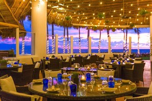 Un restaurante con mesas redondas, sillas de mimbre y vistas al océano al atardecer.