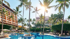 Zona de piscina en un resort, con palmeras, sillas de descanso y un edificio de varios pisos.