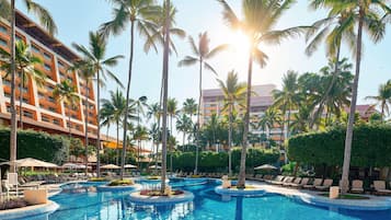 Zona de piscina en un resort, con palmeras, sillas de descanso y un edificio de varios pisos.