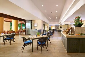 Amplia zona de comedor de hotel con numerosas mesas y sillas, una isla central con exhibición de frutas y vistas al área exterior.