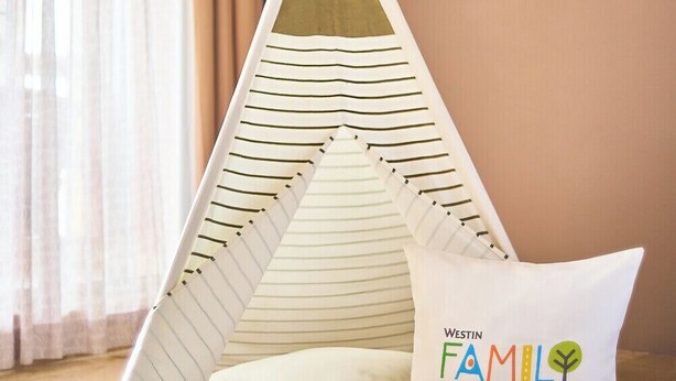 Una habitación acogedora con una tienda tipi a rayas, un cojín con la leyenda 'WESTIN FAMILY' y un frasco de vidrio, además de un folleto en el suelo.
