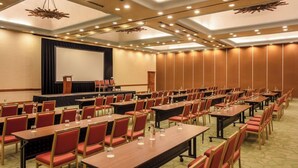 Un salón de conferencias con filas de mesas y sillas, un escenario con un atril y una pantalla grande.