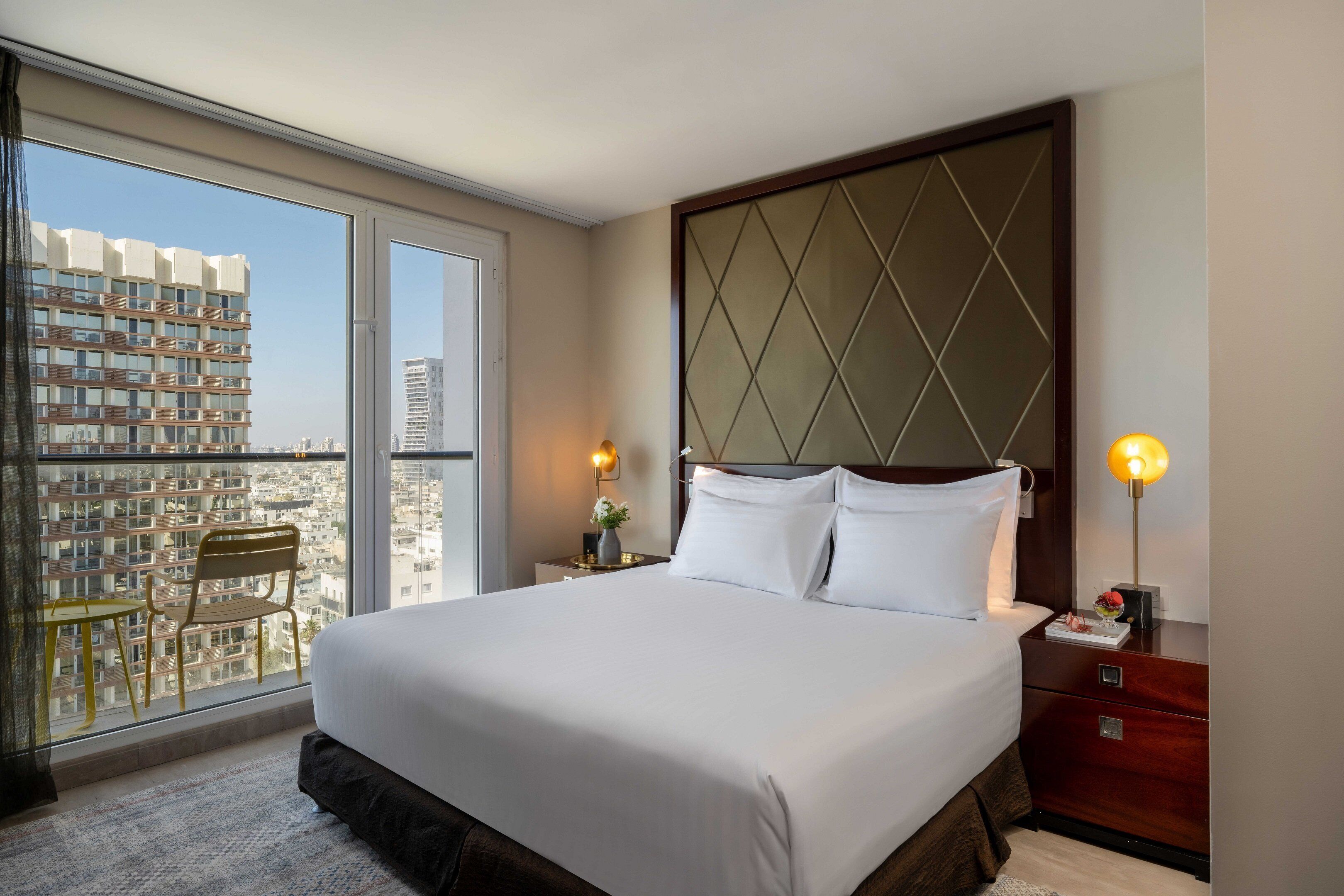 A hotel room with a large bed, a nightstand, a chair, and a view of the city through a large window.