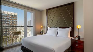 A hotel room with a large bed, a nightstand, a chair, and a view of the city through a large window.
