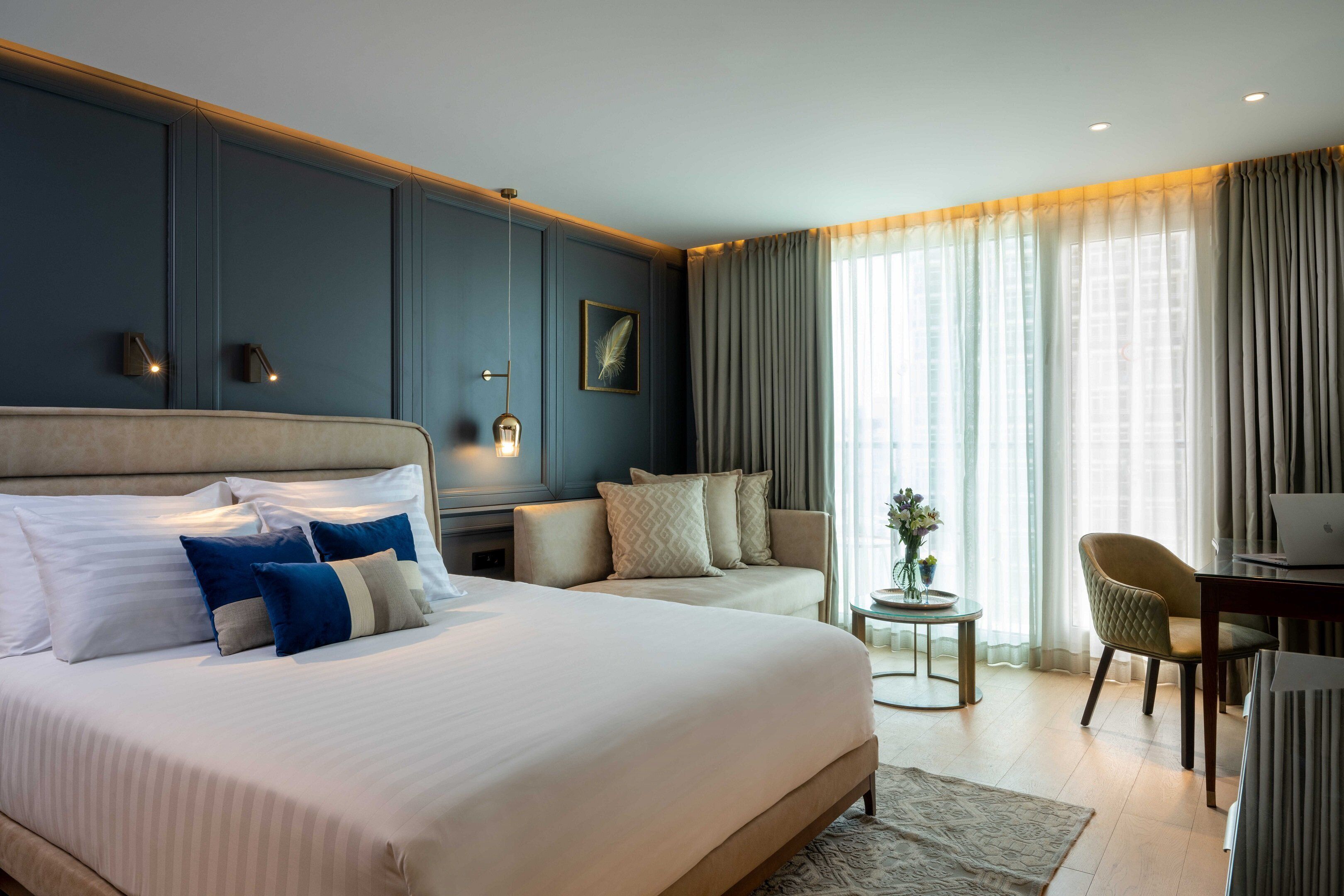 Une chambre d’hôtel avec un grand lit, un canapé, un bureau et une chaise. Il y a une fenêtre avec des rideaux et une plante sur le bureau.