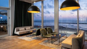A modern living room with a large window overlooking a beach, furnished with a sofa, chairs, and a coffee table. - Crowne Plaza Tel Aviv Beach by IHG (Tel Aviv)