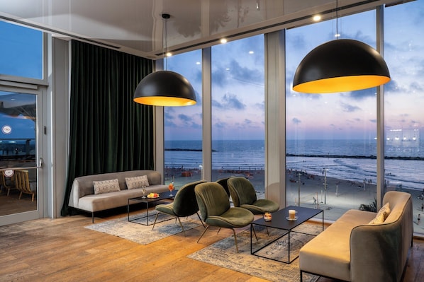 A modern living room with a large window overlooking a beach, furnished with a sofa, chairs, and a coffee table.