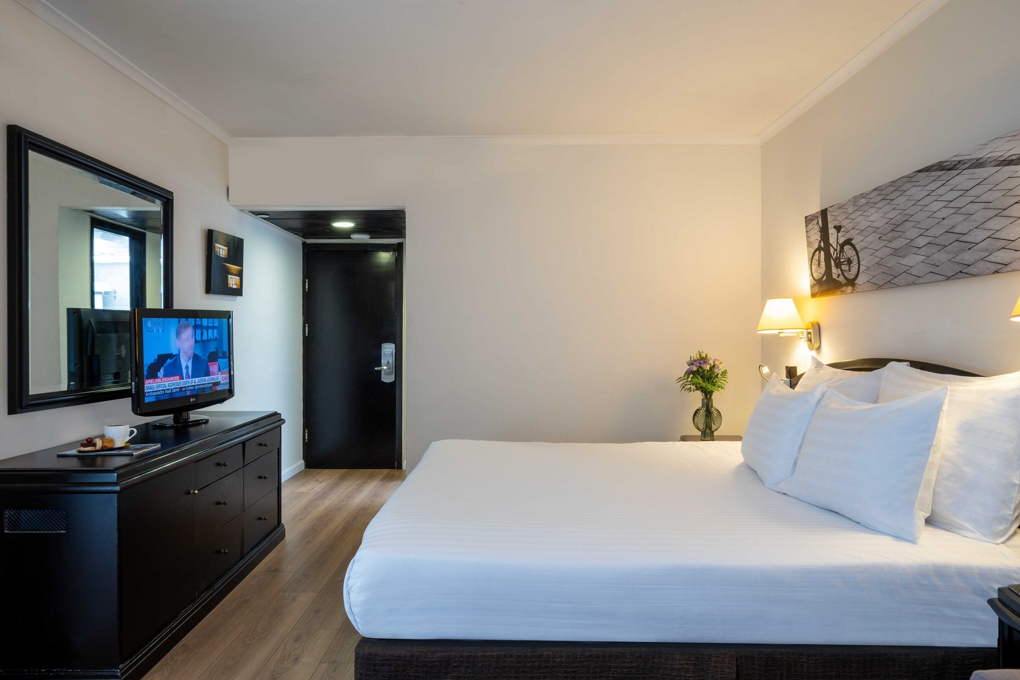 A hotel room with a large bed, a TV on a dresser, a mirror, and a wall-mounted lamp.