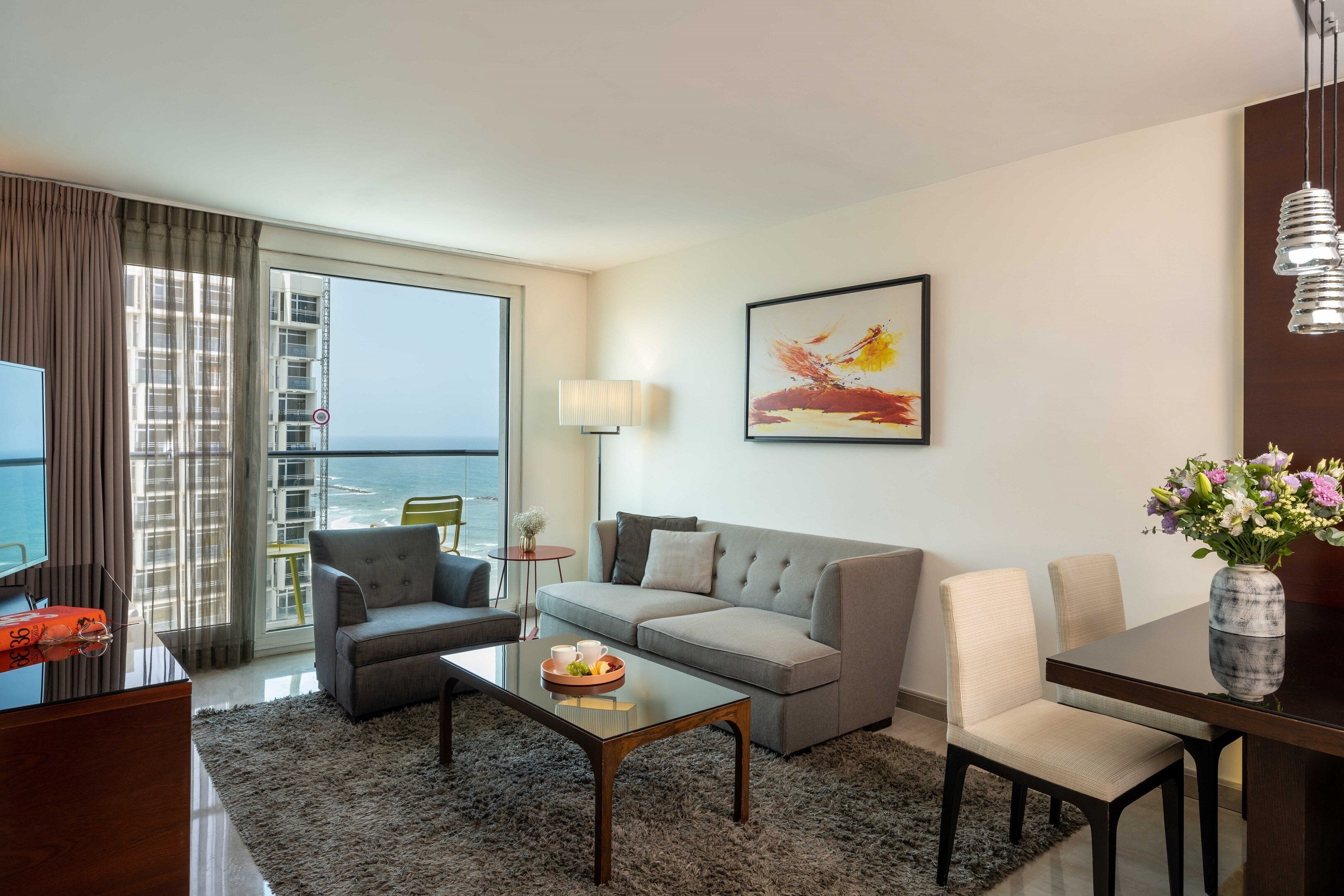 A modern hotel room with a sofa, armchairs, a coffee table, and a dining table. There is a large window with a view of the sea and a balcony.