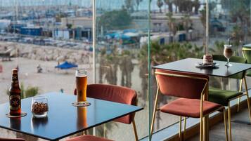 Una mesa con una botella, un vaso y un postre, con vista a una playa con barcos y sombrillas.